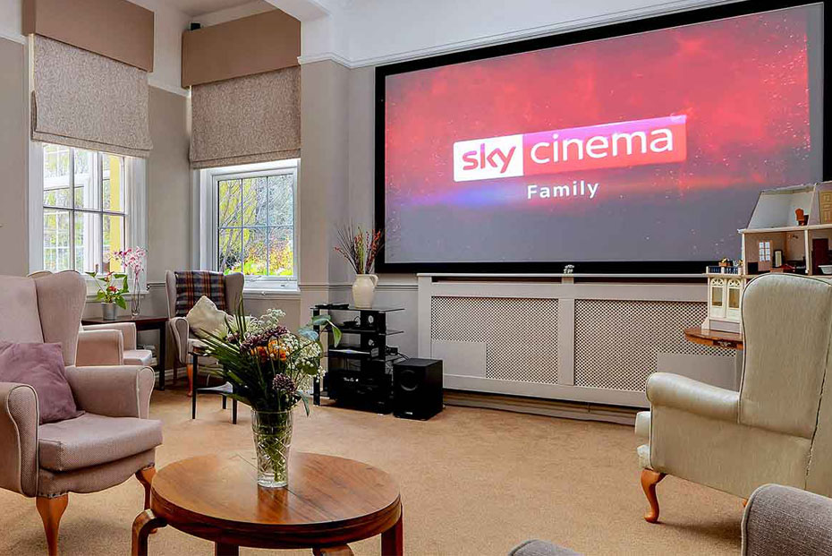 Cinema room with multiple beige armchairs facing a large screen displaying the Sky Cinema Family logo. The room has windows on one side, cream walls, and a patterned carpet.