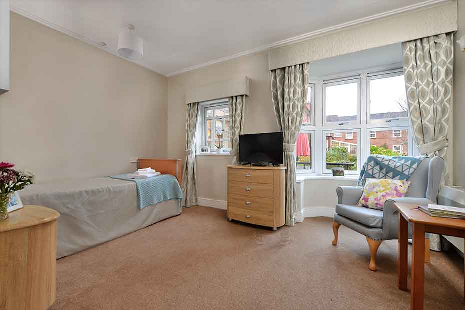 Modern styled bedroom with carpeting, featuring a single bed, a wooden chest of drawers with a TV on top, a light blue armchair with floral cushions, and patterned curtains.
