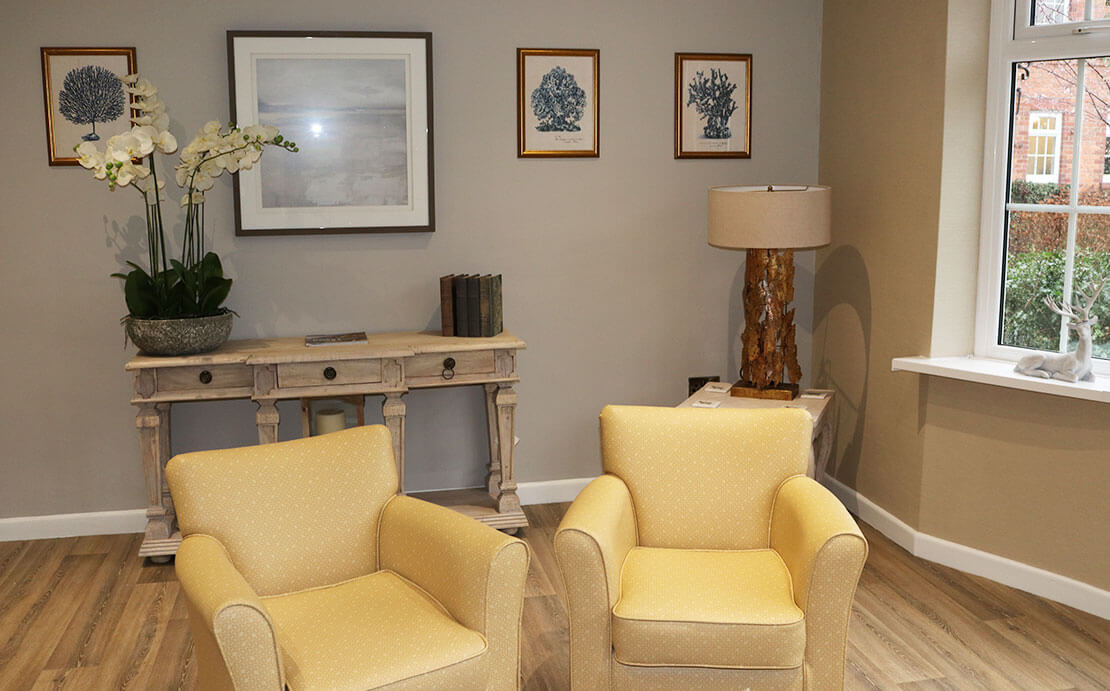 Well-lit room lounge featuring two yellow armchairs, a wooden console table adorned with decorative items, a framed painting on the wall, and three blue botanical prints.