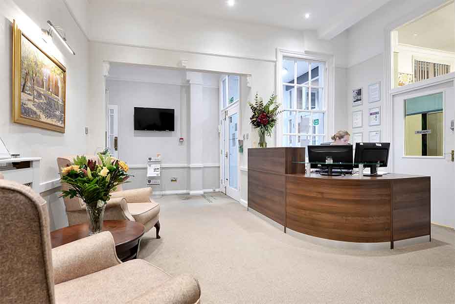 Reception area with a wooden desk with a person working on a computer, a seating area with chairs and a sofa, a wall-mounted TV, and decorative plants.