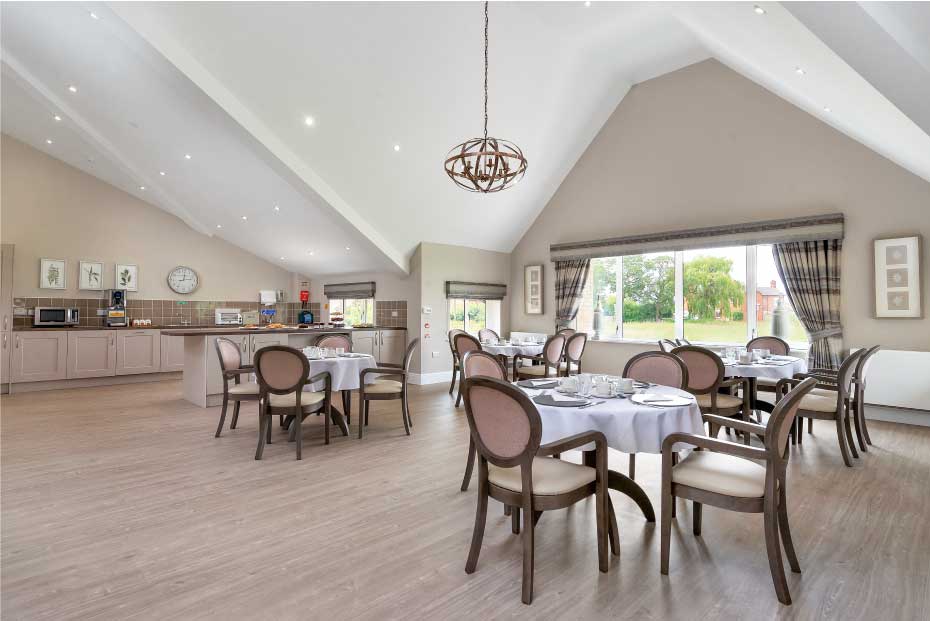 A bright, spacious dining area with round tables, cushioned chairs, a vaulted ceiling, and large windows overlooking greenery.