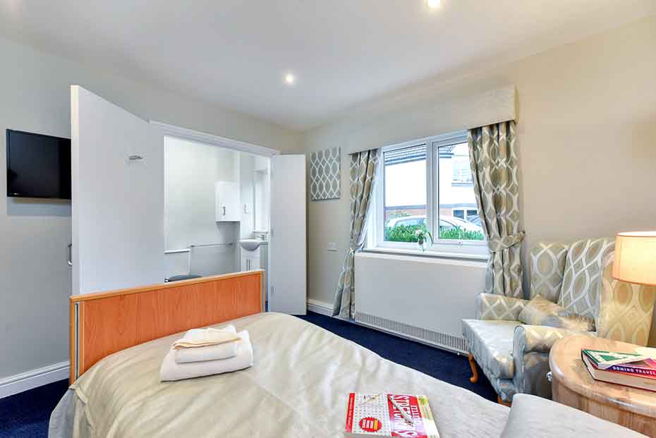 A well-lit bedroom with a double bed adorned with white bedding and a folded towel on it. There is a flat-screen TV on the wall, a small wooden desk with an open magazine, two lamps, and patterned curtains by the window.
