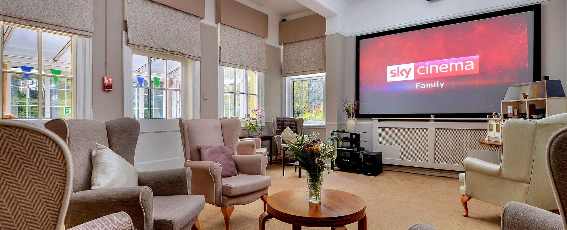 Cinema room with multiple beige armchairs facing a large screen displaying the Sky Cinema Family logo. The room has windows on one side, cream walls, and a patterned carpet.