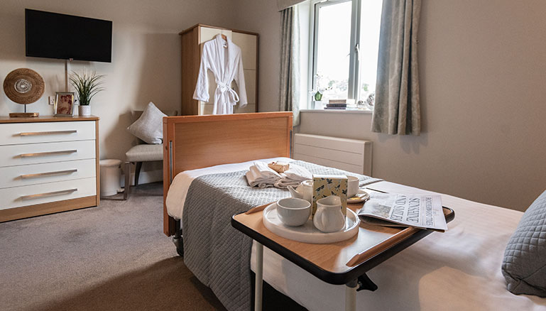 A cosy bedroom interior with natural lighting featuring a single bed, an overbed table, mounted TV, armchair and wardrobe