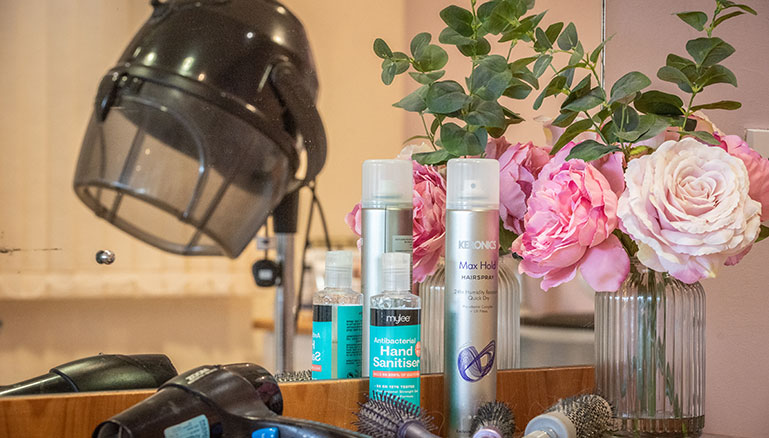 A countertop featuring a black salon hair dryer helmet, hairspray, hairbrushes and combs, and a vase with pink roses